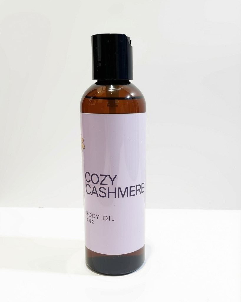 An amber glass bottle of Cozy Cashmere body oil with a black cap and a pale lavender label, set against a plain white background.